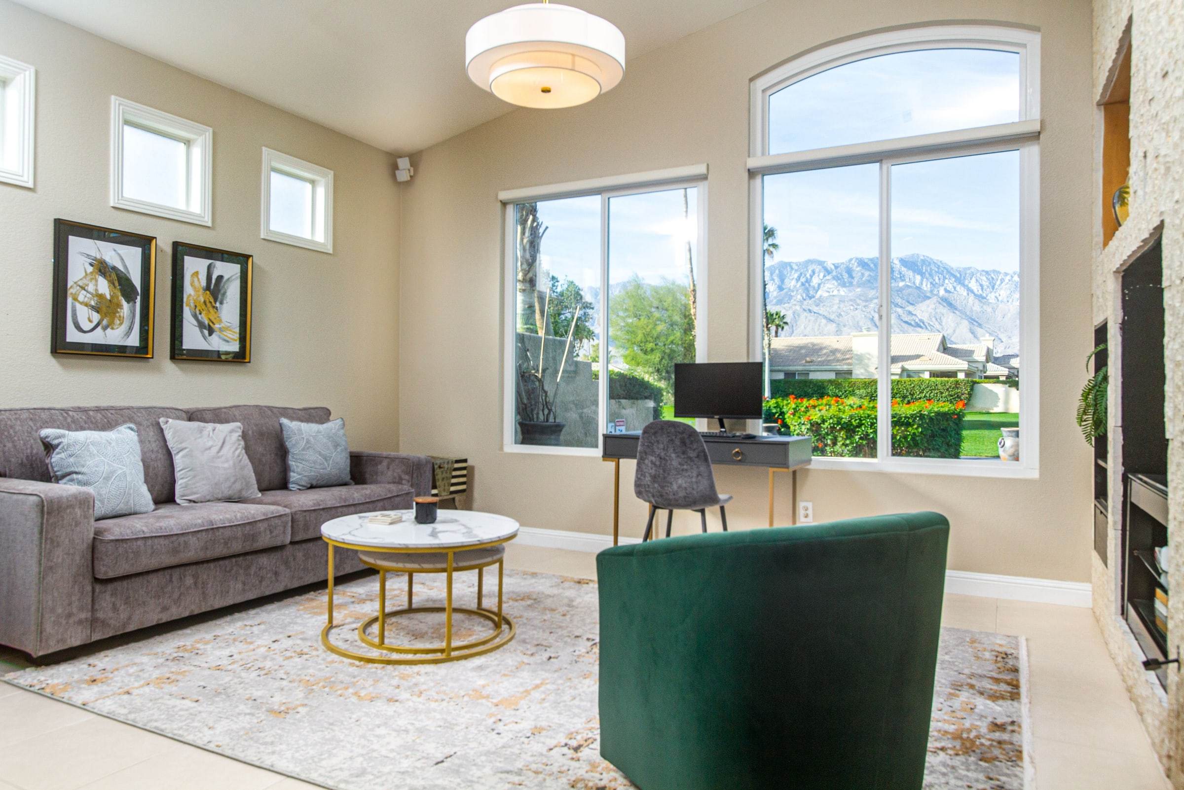 Den with a gray sofa, a green velvet armchair, marble side tables, a desk and an arched window