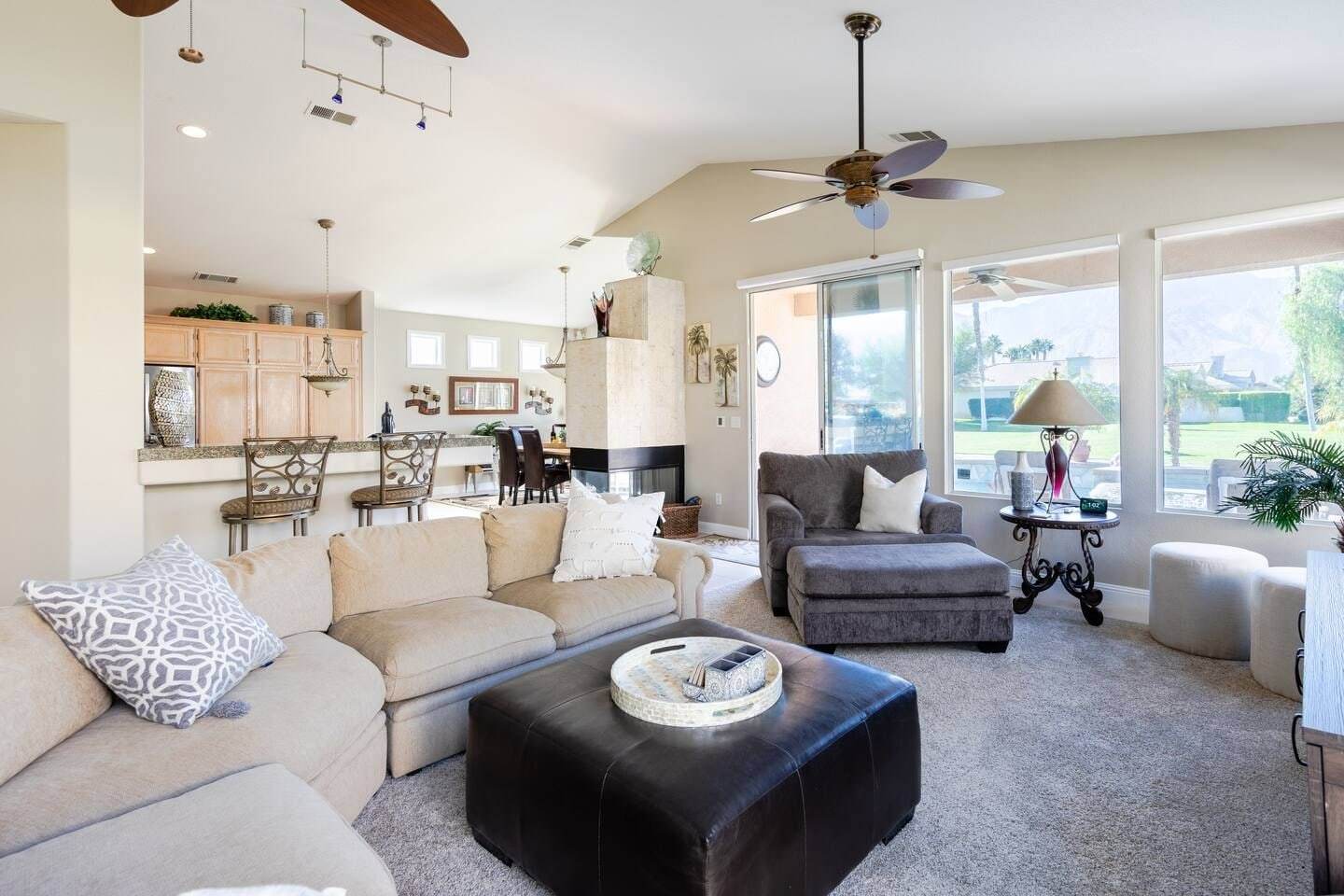 Living room with a cream sectional and leather ottoman, open to the kitchen bar and dining table, with windows onto the pool