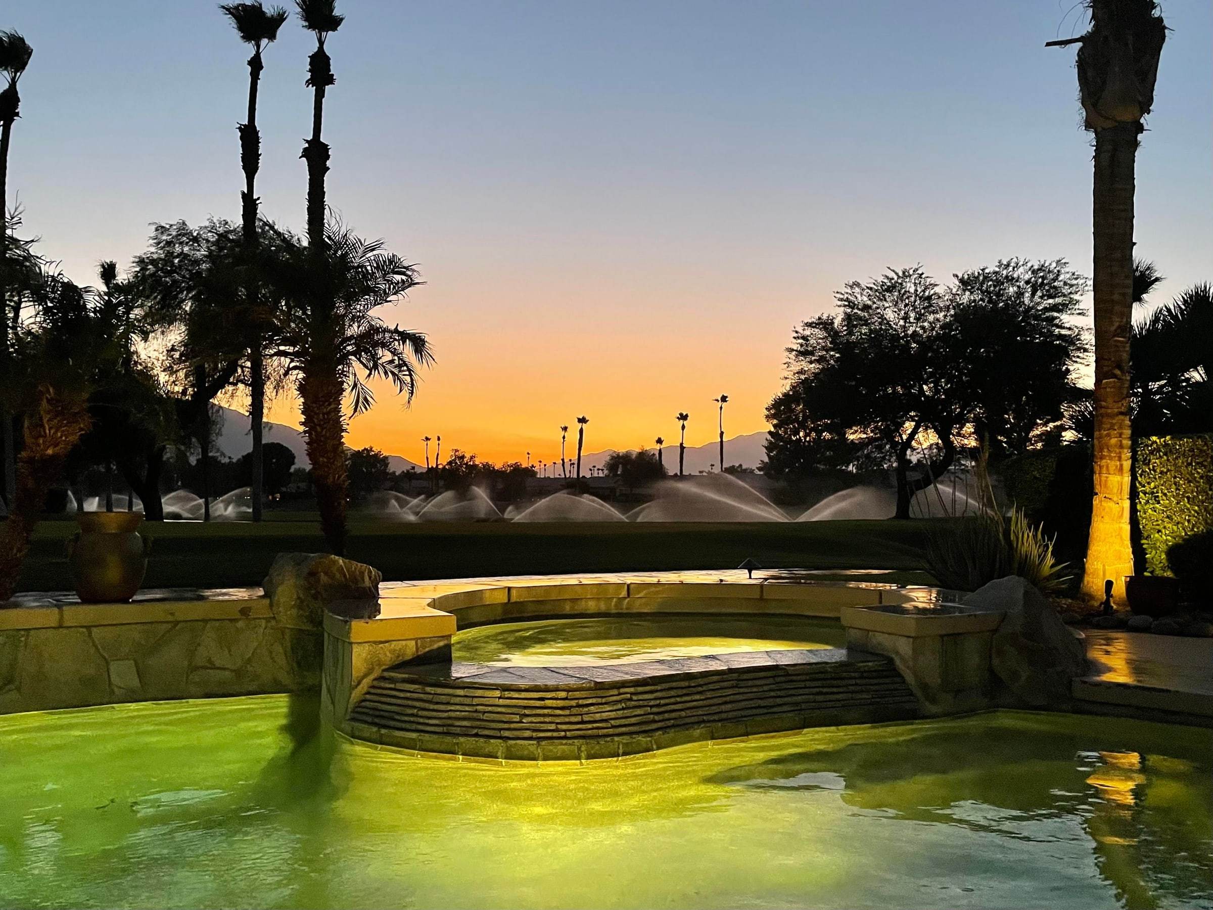 Pool and spa lit up at dusk, with sprinklers on the golf course and an orange sky behind the palms